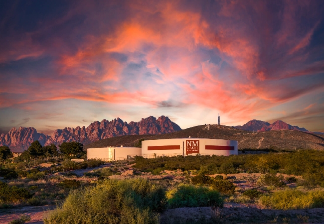 Two sets of buildings have the NMSU logo, situated outside campus an at the foothills of the mountains during sunset.