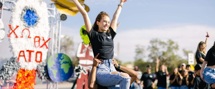 An NMSU student being held on someone's shoulders in front of a parade float