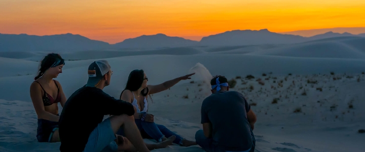 A group of college students are in the dunes at dusk, one person is pointing to the horizon.