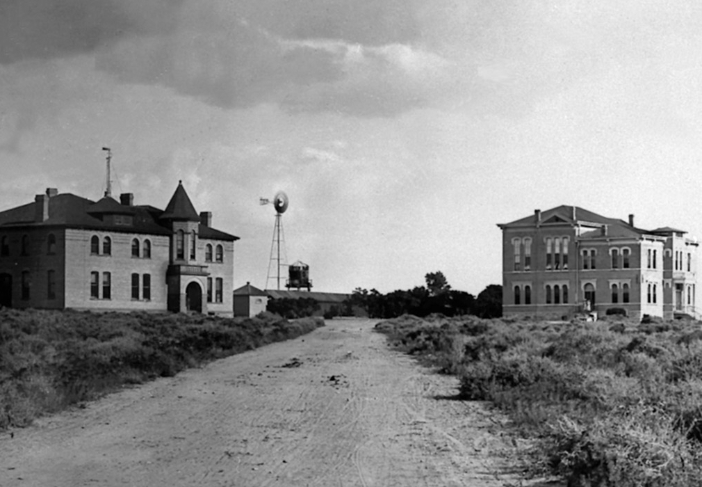 A historic black and white photo of the early stages of construction on NMSU campus, showing two large buildings and a farm windmill in the distance.